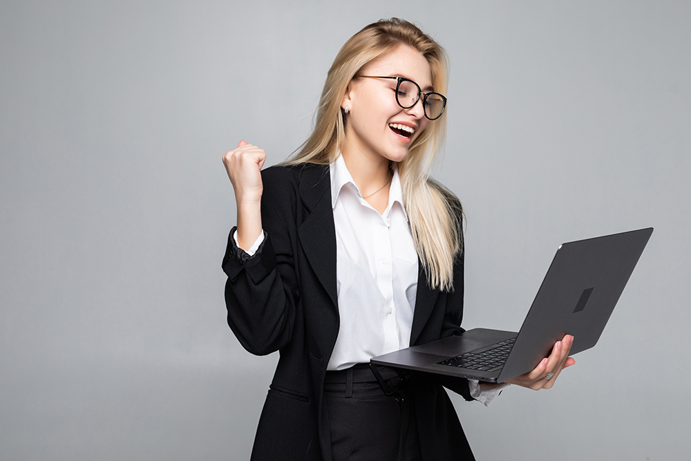 Portrait Of A Young Happy Business Woman With A Laptop With Win Gesture Over Gray Background